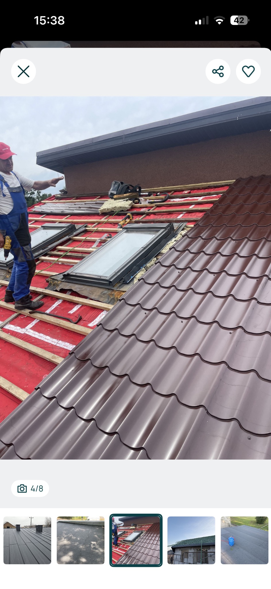 Montaż brązowej blachodachówki na dachu z oknami połaciowymi. Widoczny dekarz w kombinezonie i czerwonej czapce podczas pracy. Częściowo ułożone pokrycie.