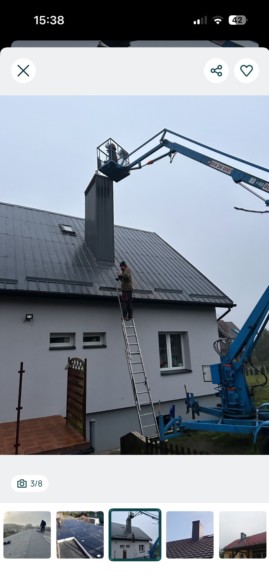 Pracownik na drabinie przy dachu z blachy, obok podnośnik koszowy. Szary dach i komin po renowacji. Dom z jasną elewacją w tle.