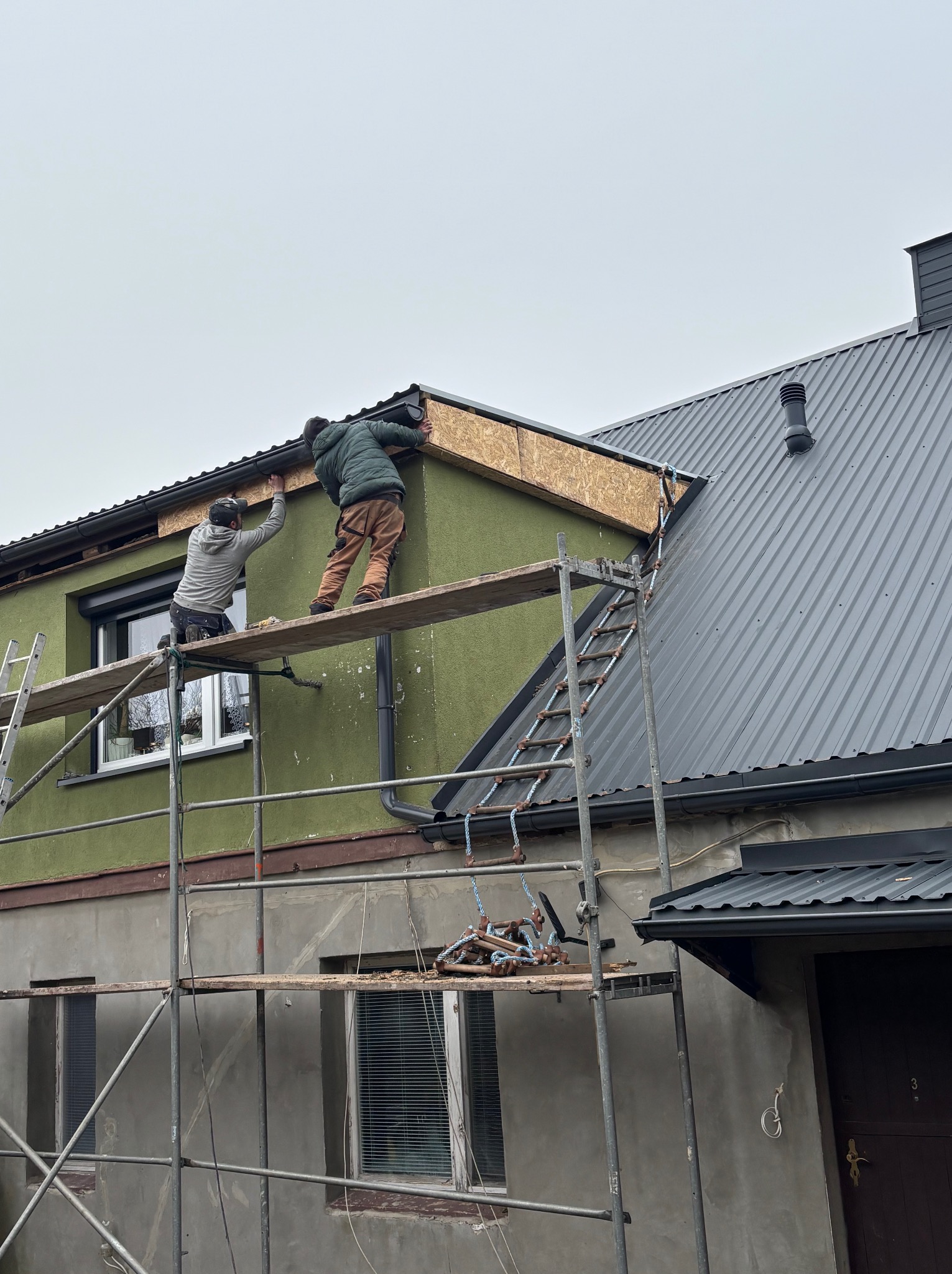 Dwóch monterów na rusztowaniu mocuje elementy wykończeniowe dachu przy zielonej elewacji domu z szarą blachodachówką. Widoczne narzędzia i drabina.