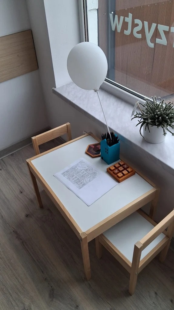 Dziecięcy stół z krzesłem, rysunek osiedla, gry edukacyjne i biały balon przy oknie. Ujęcie z góry, neutralne oświetlenie.