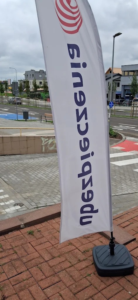 Biały, pionowy baner reklamowy z logo i tekstem 'Ubezpieczenia' ustawiony na chodniku z kostki brukowej, na tle ulicy i budynków w pochmurny dzień.