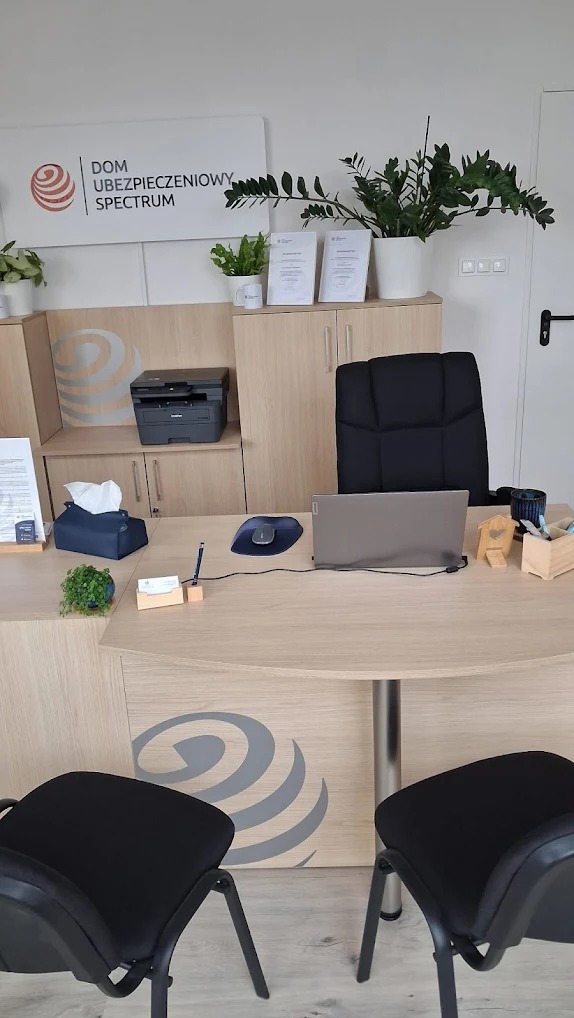 Biuro ubezpieczeniowe z biurkiem, laptopem, krzesłami dla klientów i logo firmy 'Dom Ubezpieczeniowy Spectrum' na ścianie. Wnętrze jasne, z roślinami doniczkowymi.