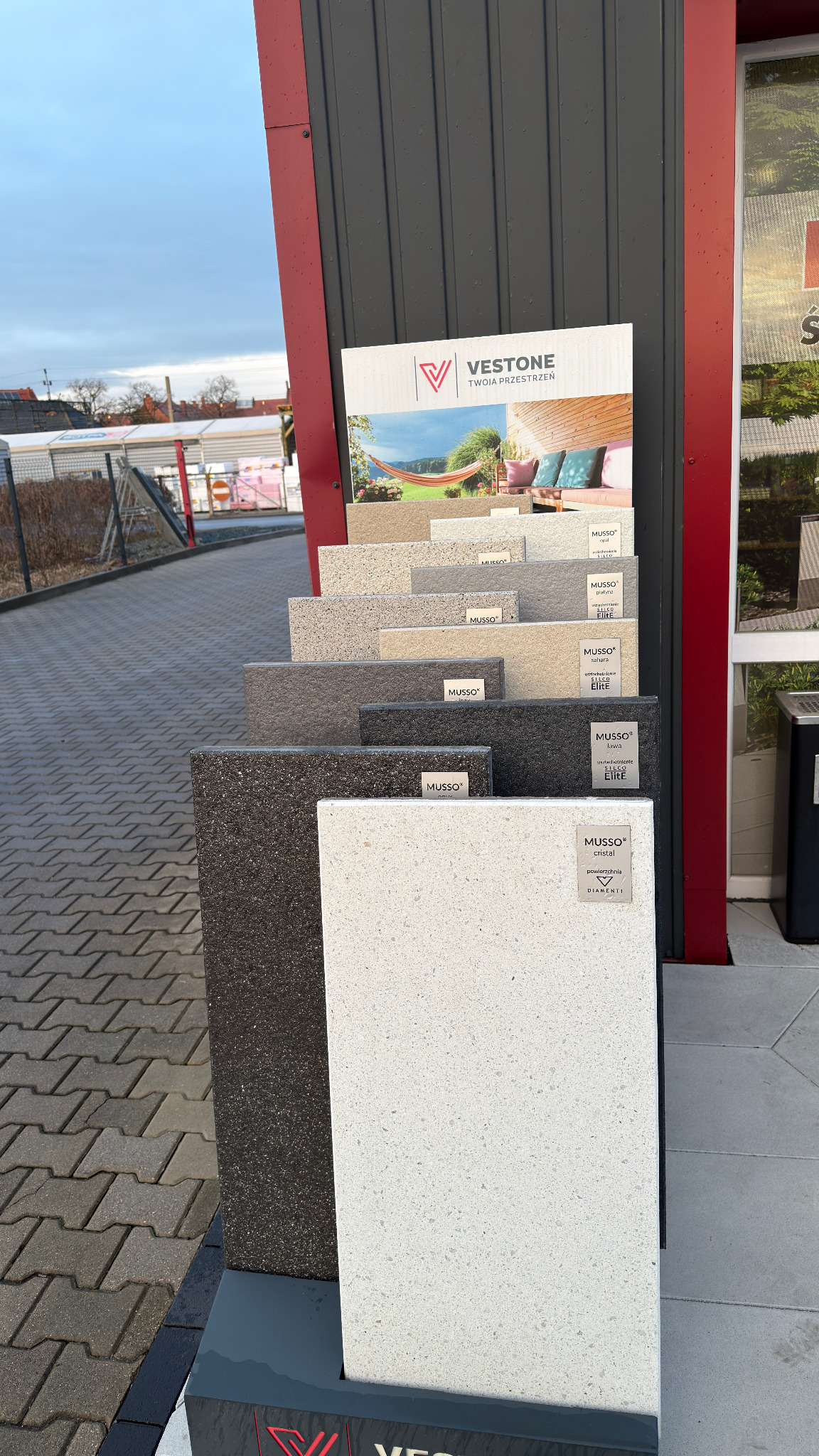 Ekspozycja próbek płyt Vestone Musso w Wołowie. Różne kolory i faktury, od jasnych po ciemne, prezentowane na stojaku przed budynkiem. Widoczna kostka brukowa.