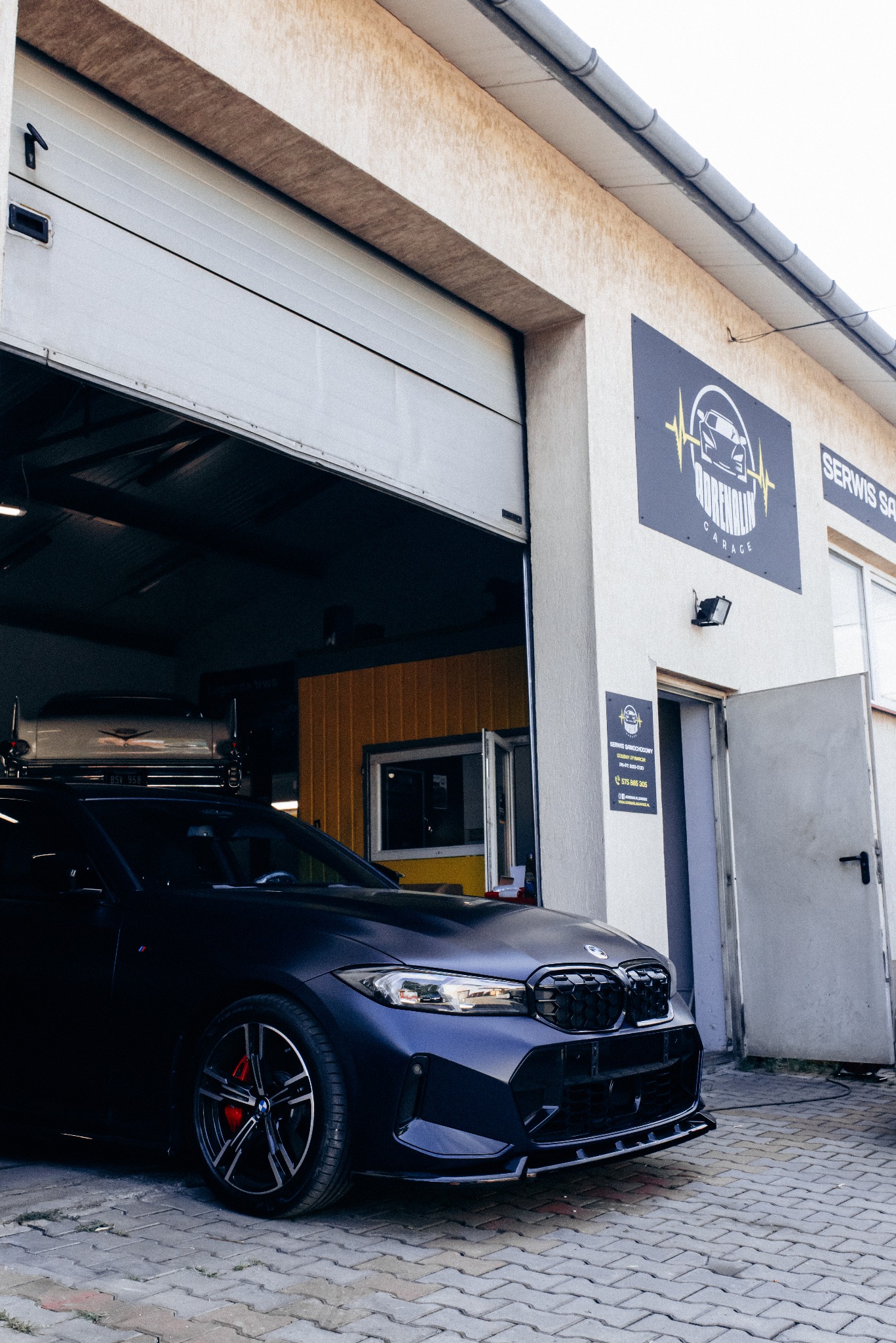 Granatowy, zmodyfikowany samochód BMW przed otwartym garażem warsztatu z logo 'Adrenalin Garage' i informacją 'Serwis Samochodowy'. Czerwone zaciski hamulcowe.