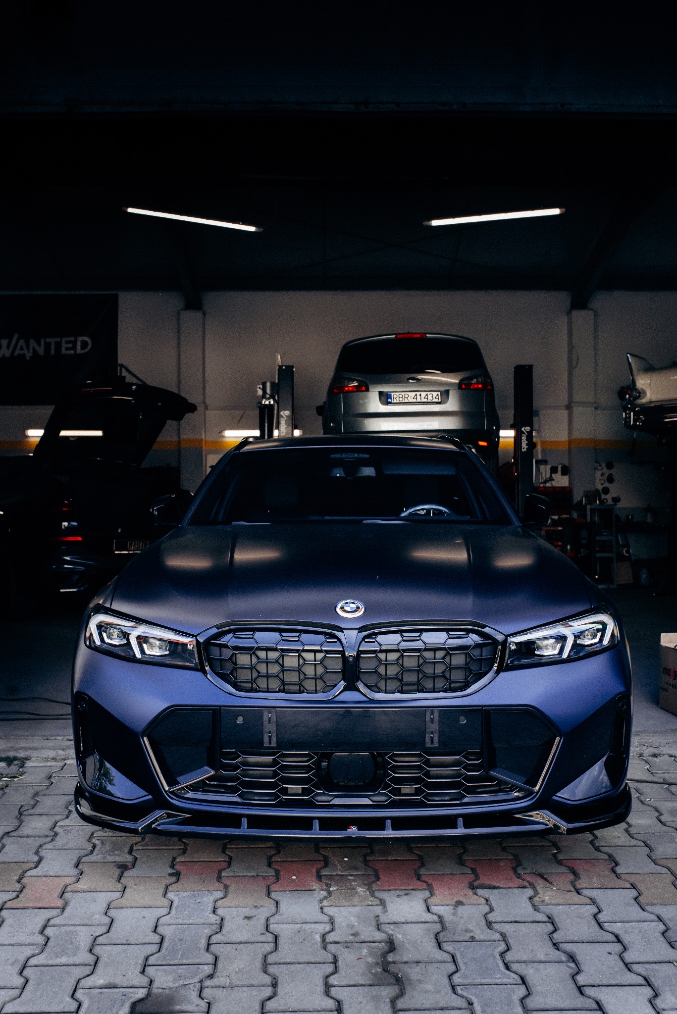 Granatowy, matowy BMW serii 3 (G20) po tuningu, z agresywnym body kitem, na tle warsztatu z podnośnikami i innymi autami. Ujęcie z przodu, na kostce brukowej.