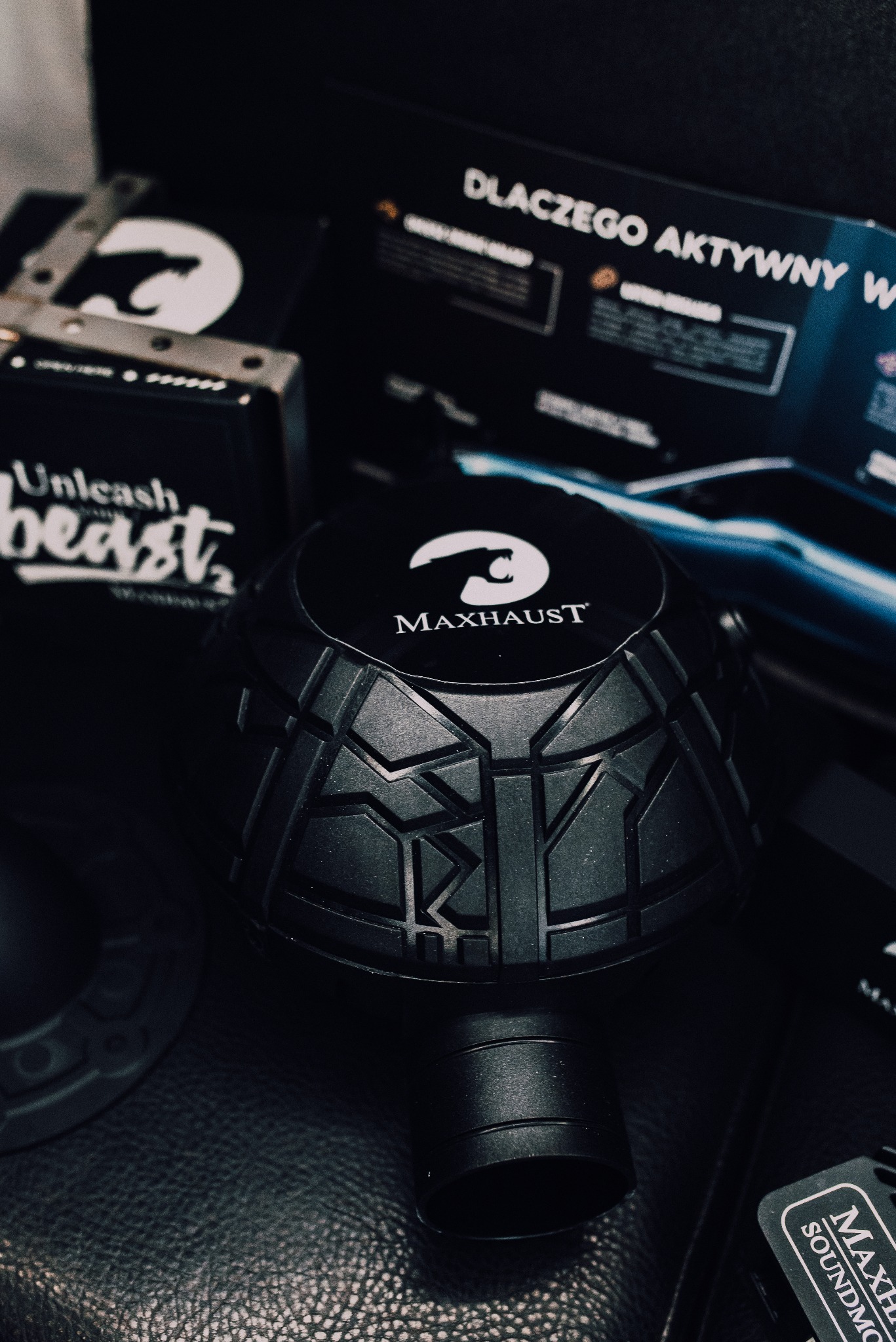 Czarny, okrągły dyfuzor akustyczny Maxhaust z logo, umieszczony na skórzanym tle, otoczony akcesoriami i materiałami reklamowymi marki. Nastrojowe oświetlenie.