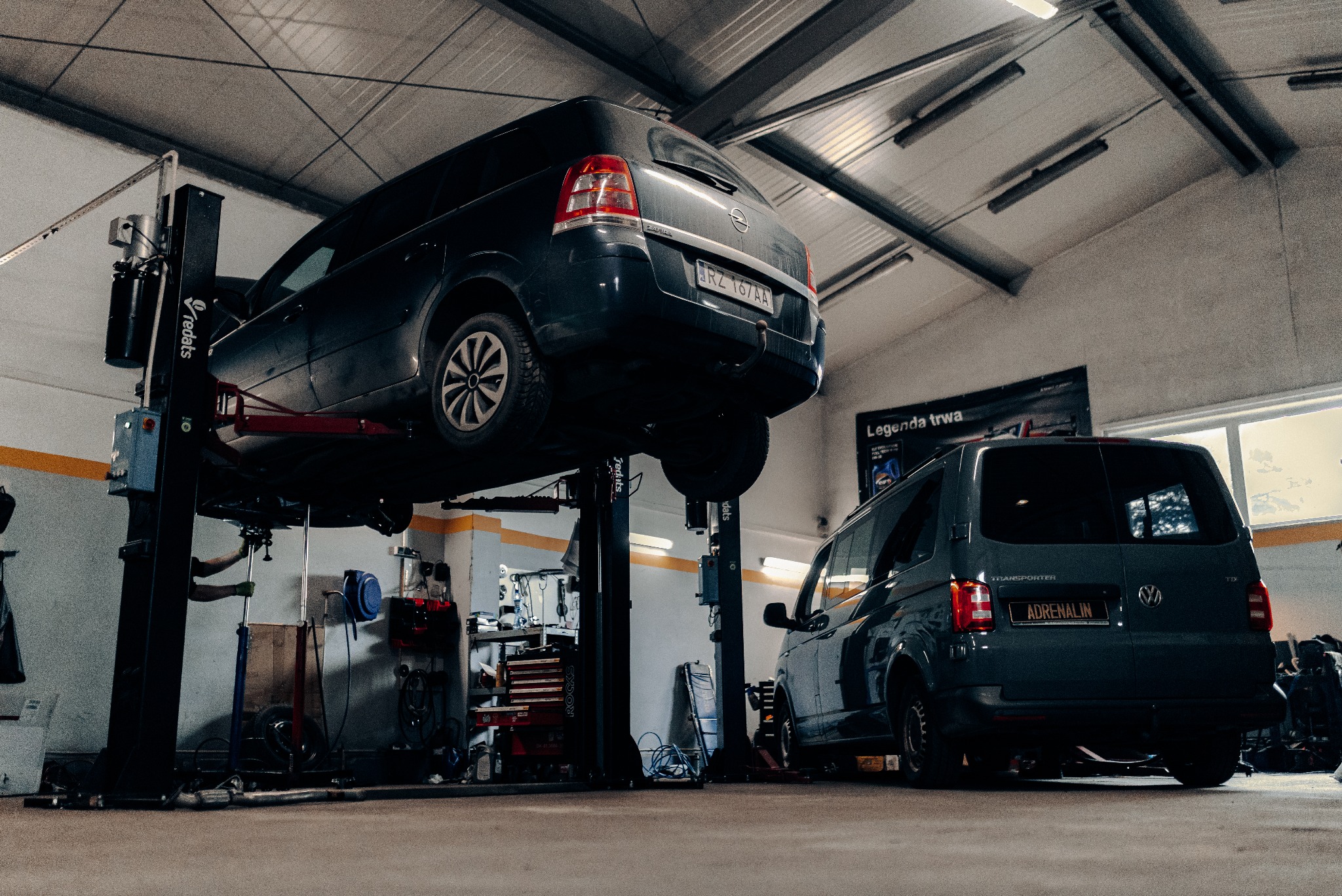 Samochód Opel Zafira na podnośniku w warsztacie, obok zaparkowany Volkswagen Transporter. Widoczne narzędzia i wyposażenie warsztatowe.