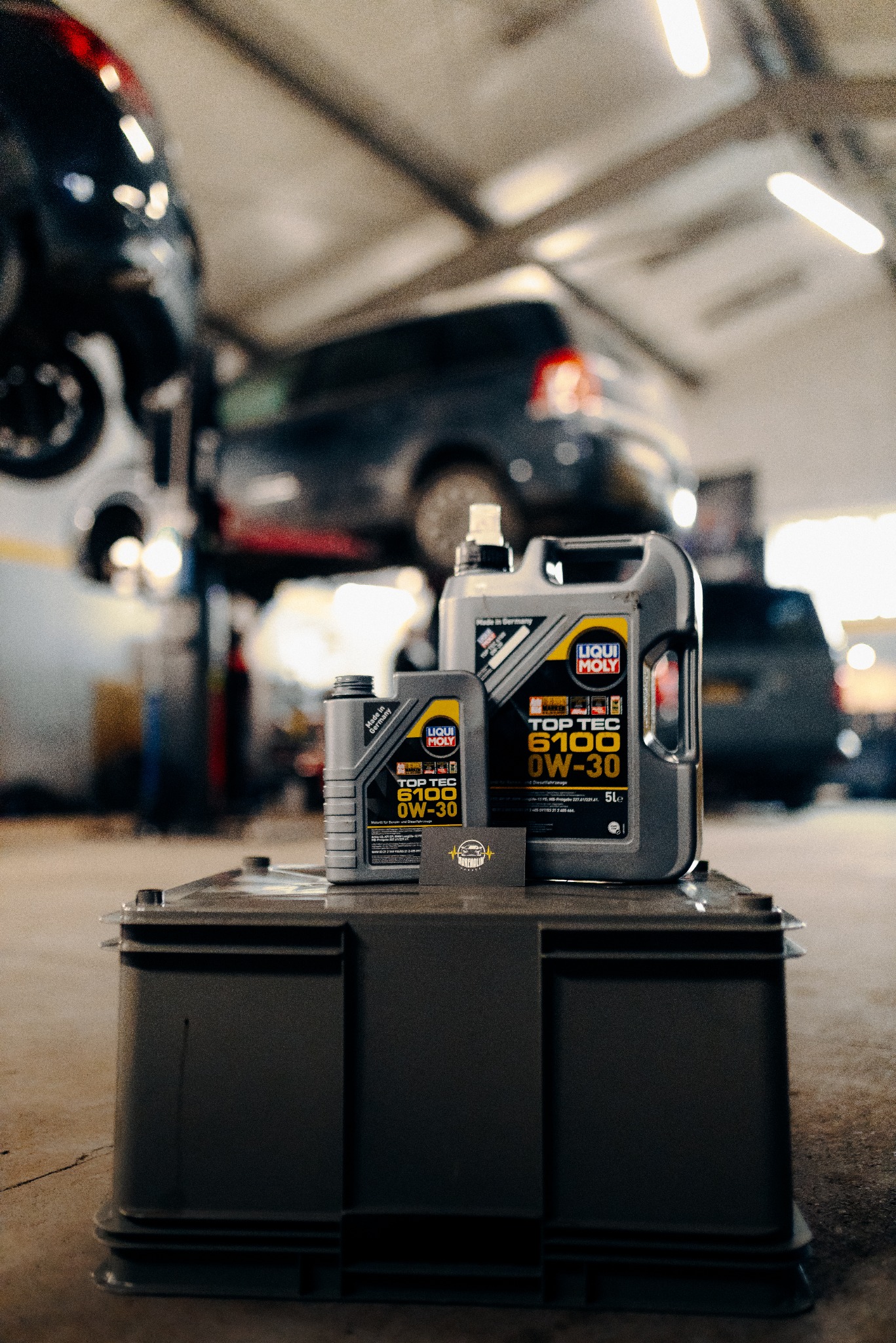 Olej silnikowy Liqui Moly 0W-30 na tle warsztatu z podnośnikiem i samochodami. Widoczna wizytówka firmy. Profesjonalne oświetlenie, ciepłe barwy.