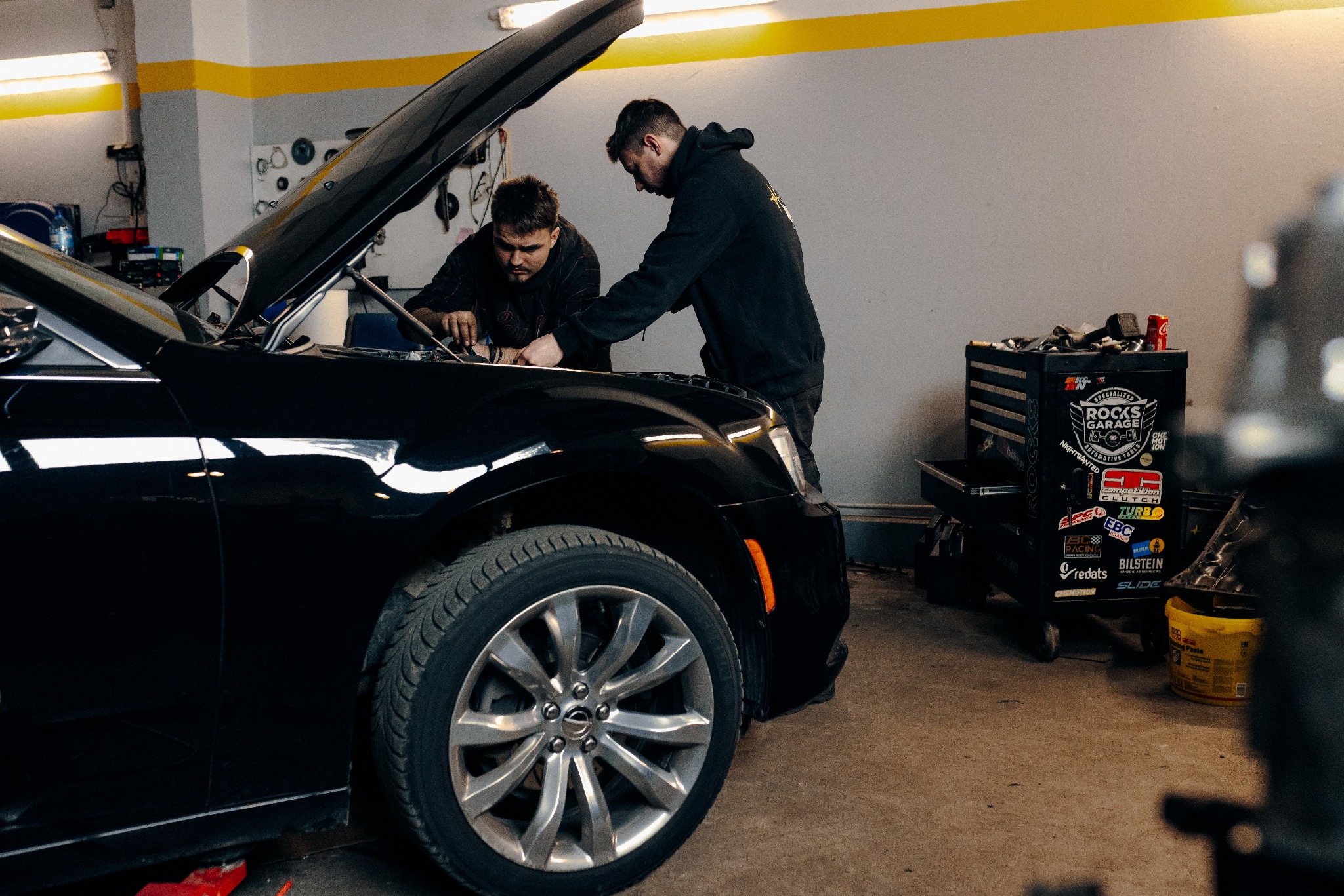 Dwóch mechaników pochylonych nad silnikiem czarnego auta z otwartą maską w warsztacie. W tle wózek z narzędziami z naklejkami.