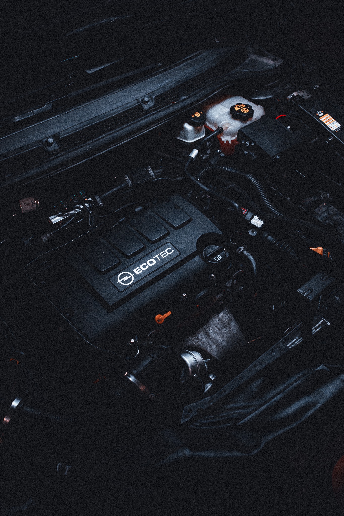 Silnik Opel ECOTEC w ciemnym ujęciu z góry. Widoczne przewody, zbiorniki płynów i osłona silnika. Ciemna tonacja zdjęcia.