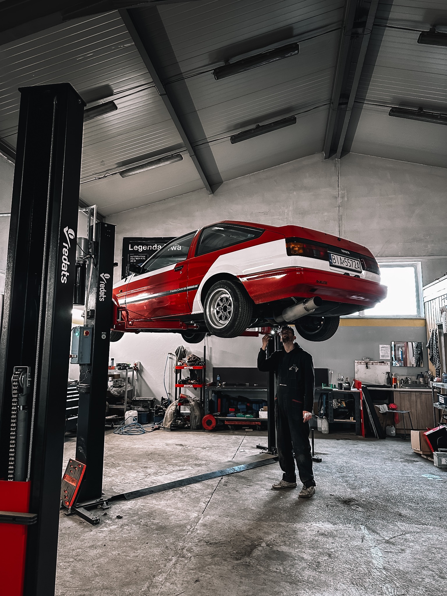 Czerwono-biała Toyota AE86 na podnośniku w warsztacie. Mechanik w kombinezonie sprawdza podwozie. Widoczne narzędzia i akcesoria warsztatowe.