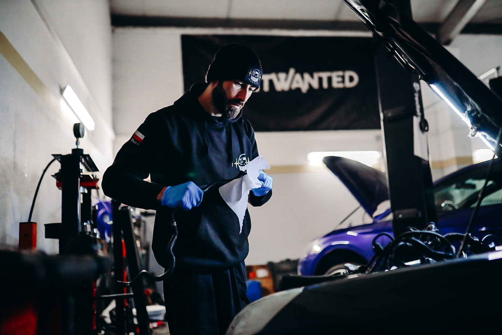Mężczyzna w czarnej bluzie i niebieskich rękawiczkach, z papierem w dłoniach, pochyla się nad silnikiem samochodu z otwartą maską w warsztacie.