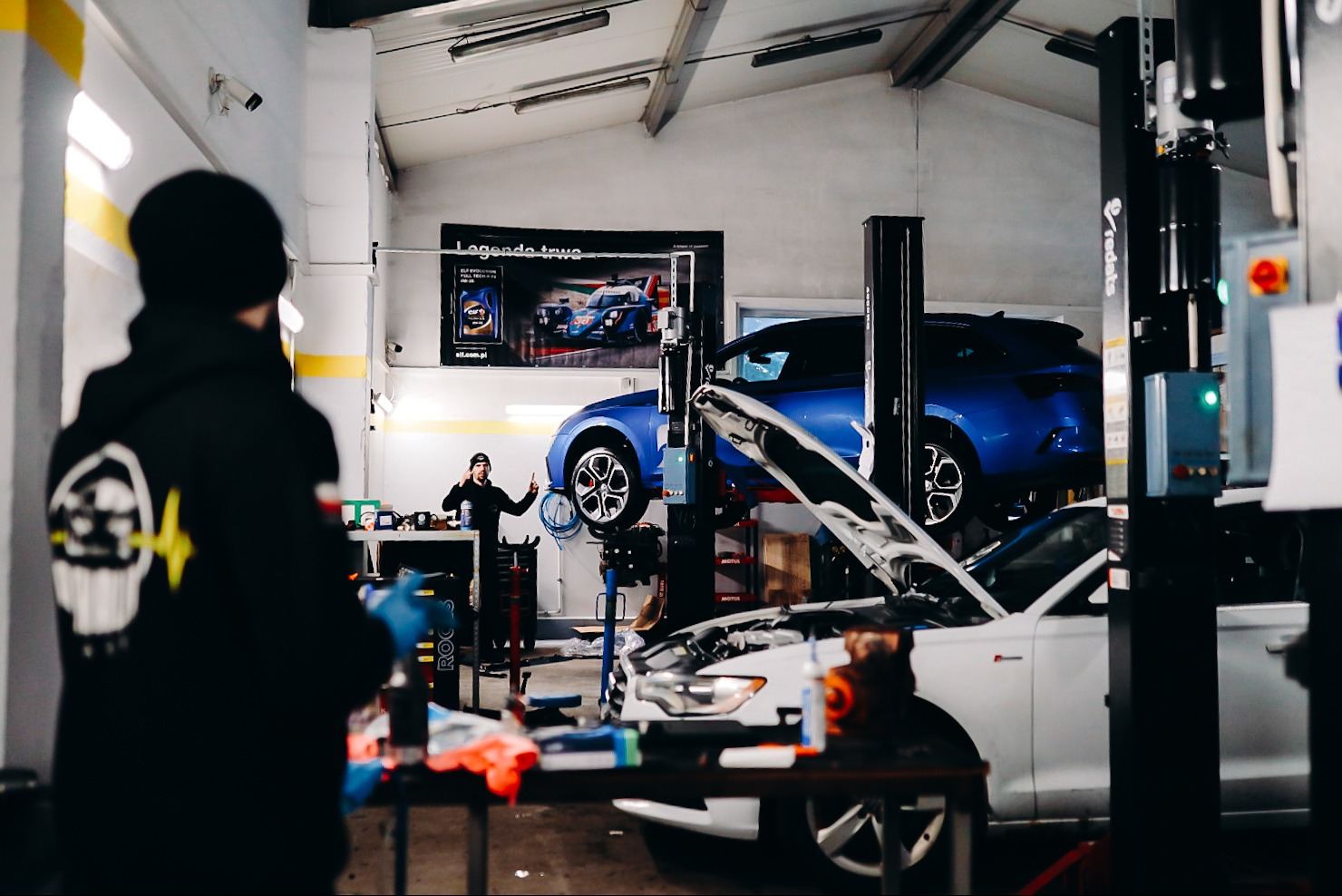 Wnętrze warsztatu: białe auto z otwartą maską na pierwszym planie, niebieskie auto na podnośniku w tle, mechanik w tle rozmawia przez telefon.