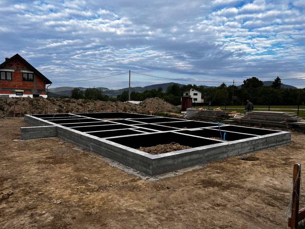 Zalane fundamenty pod budynek jednorodzinny w Podegrodziu. Widoczne szalunki i przygotowane zbrojenie. W tle dom w stanie surowym otwartym i malowniczy krajobraz.