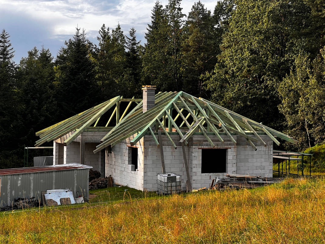 Surowy stan domu z pustaków z drewnianą konstrukcją dachu na tle lasu. Widoczne materiały budowlane i trawa. Ujęcie w słoneczny dzień.