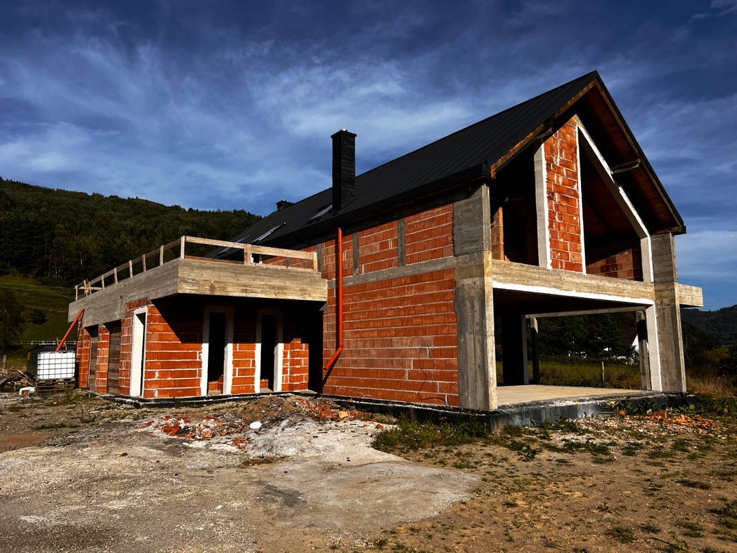 Surowy stan budynku z czerwonej cegły i betonowych elementów konstrukcyjnych, czarny dach, widok na górzysty krajobraz w tle, wczesny etap budowy.
