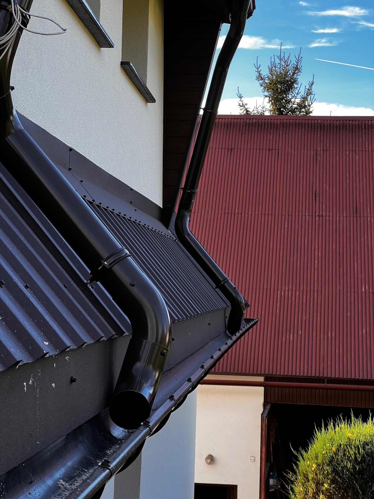 Czarny system rynnowy na dachu z blachy trapezowej, widok z dołu, na tle czerwonego dachu i błękitnego nieba z śladem samolotu. Detal montażu rynien.