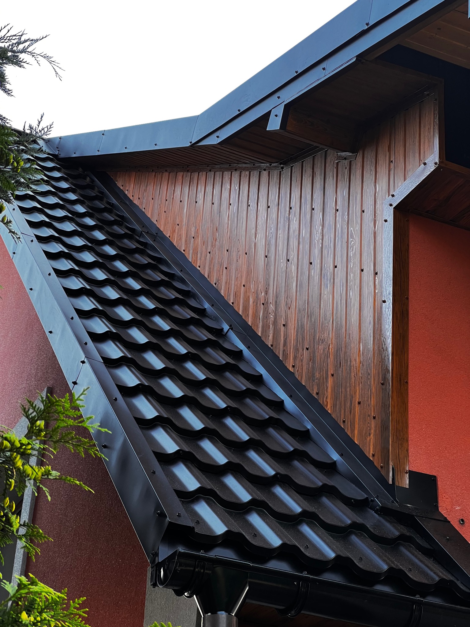 Czarny dach z blachy trapezowej, widok z dołu, z rynną. Elewacja z drewnianą boazerią i fragmentem ściany w kolorze ceglastym. Ujęcie z dołu, zieleń w kadrze.