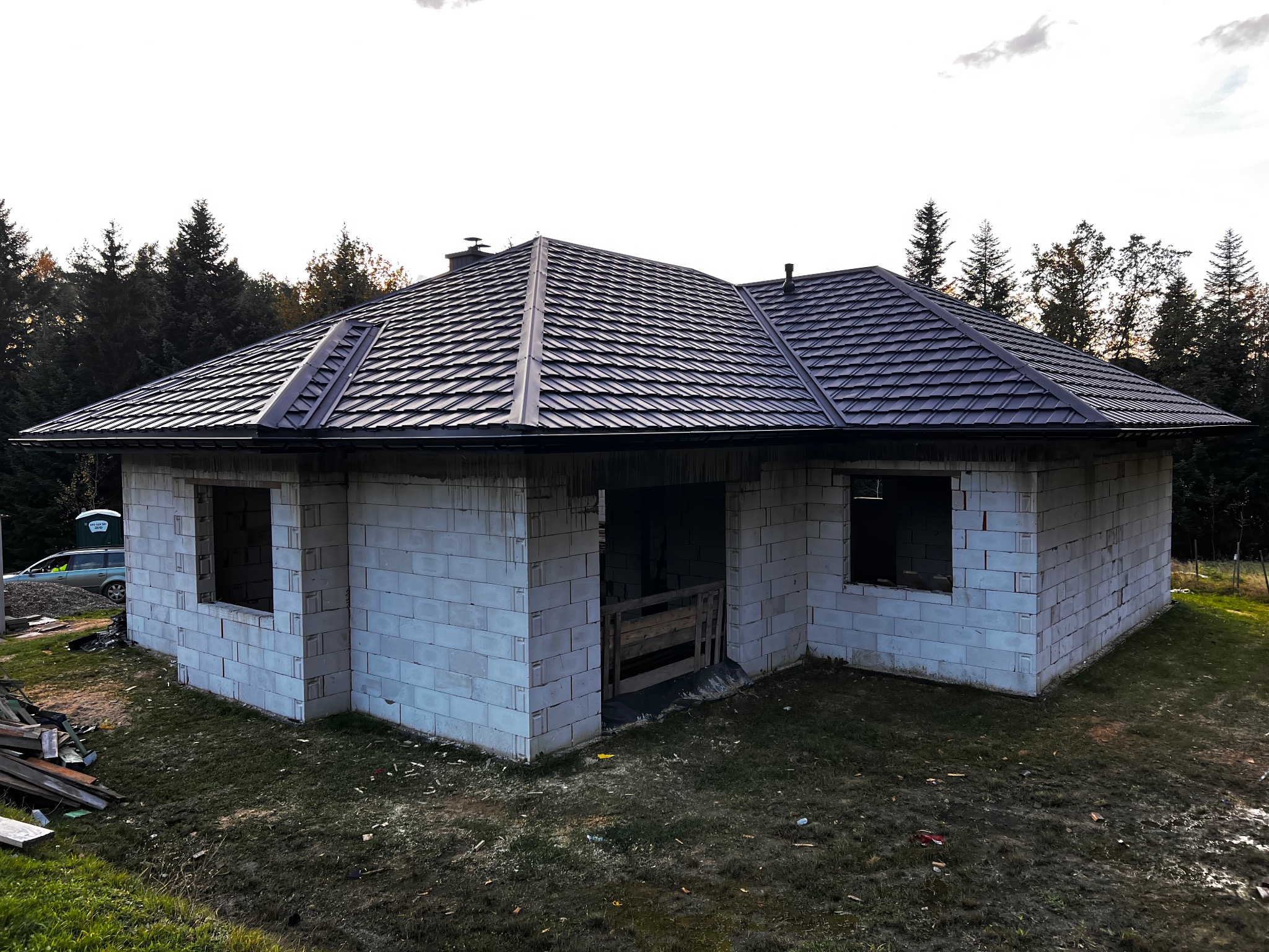 Niedokończony dom z ciemnym dachem z dachówki, widoczne białe ściany z betonu komórkowego i otwory okienne. W tle drzewa i niebo.