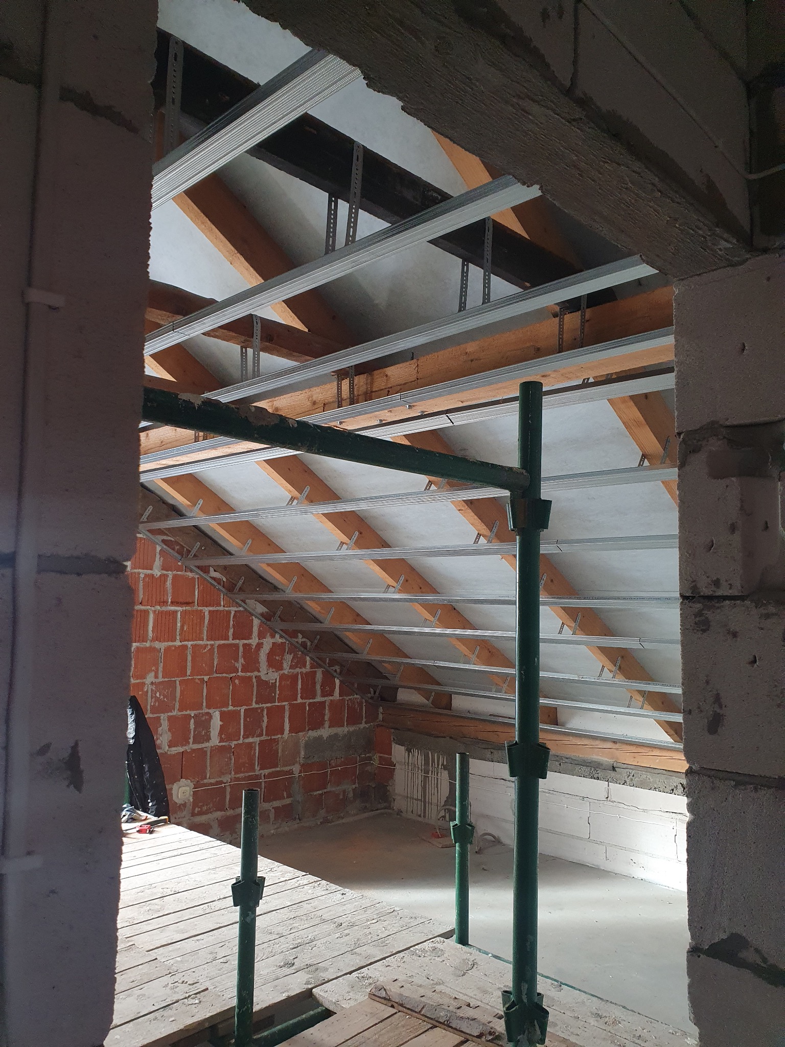 Sufit podwieszany z metalową konstrukcją i płytami gipsowymi w trakcie montażu, widok z rusztowania. Fragment ściany z cegły i bloczków.