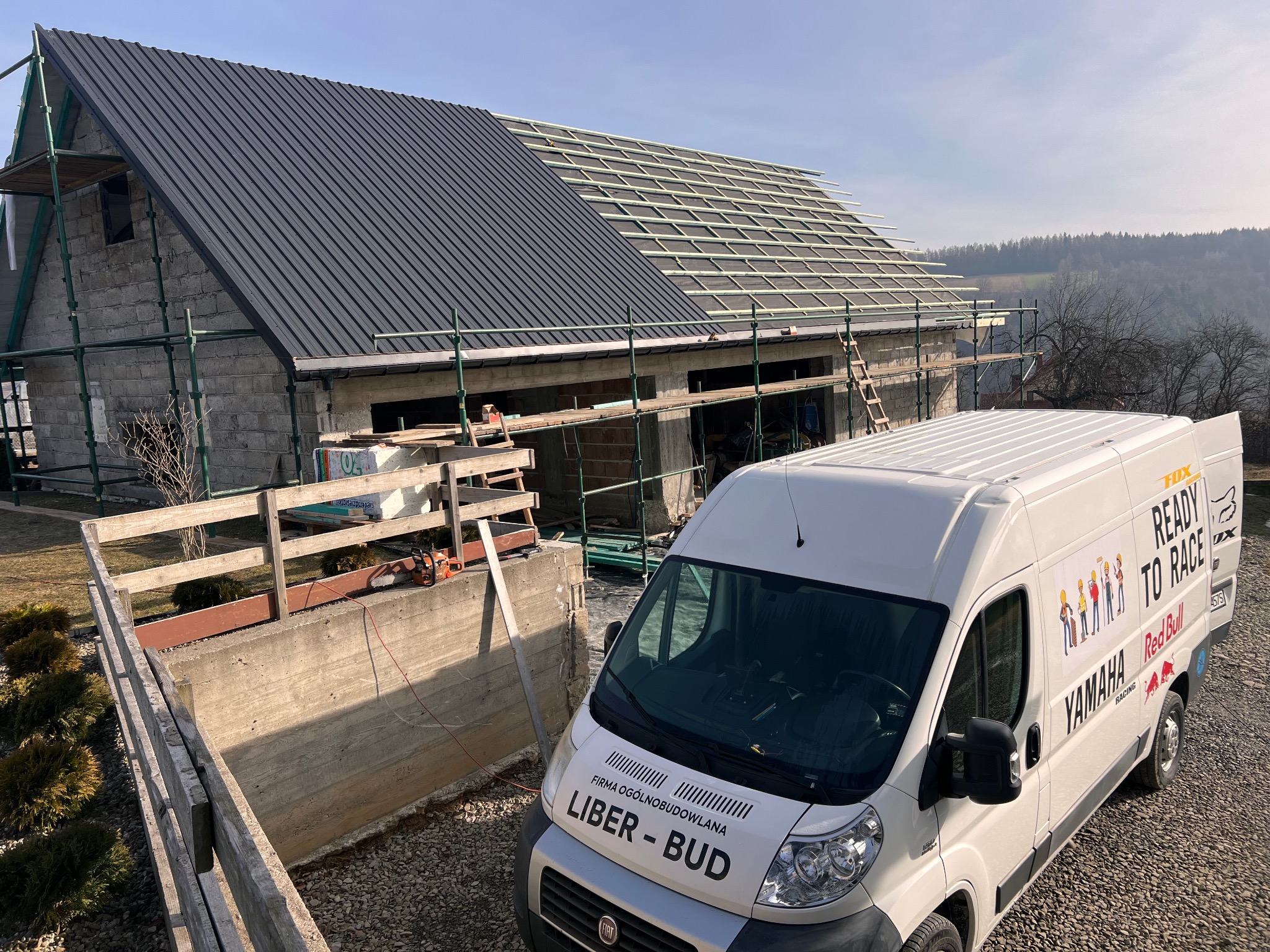 Budowa domu jednorodzinnego w trakcie montażu dachu z blachy na rąbek stojący. Część dachu pokryta, druga w trakcie. Na pierwszym planie samochód firmy budowlanej.