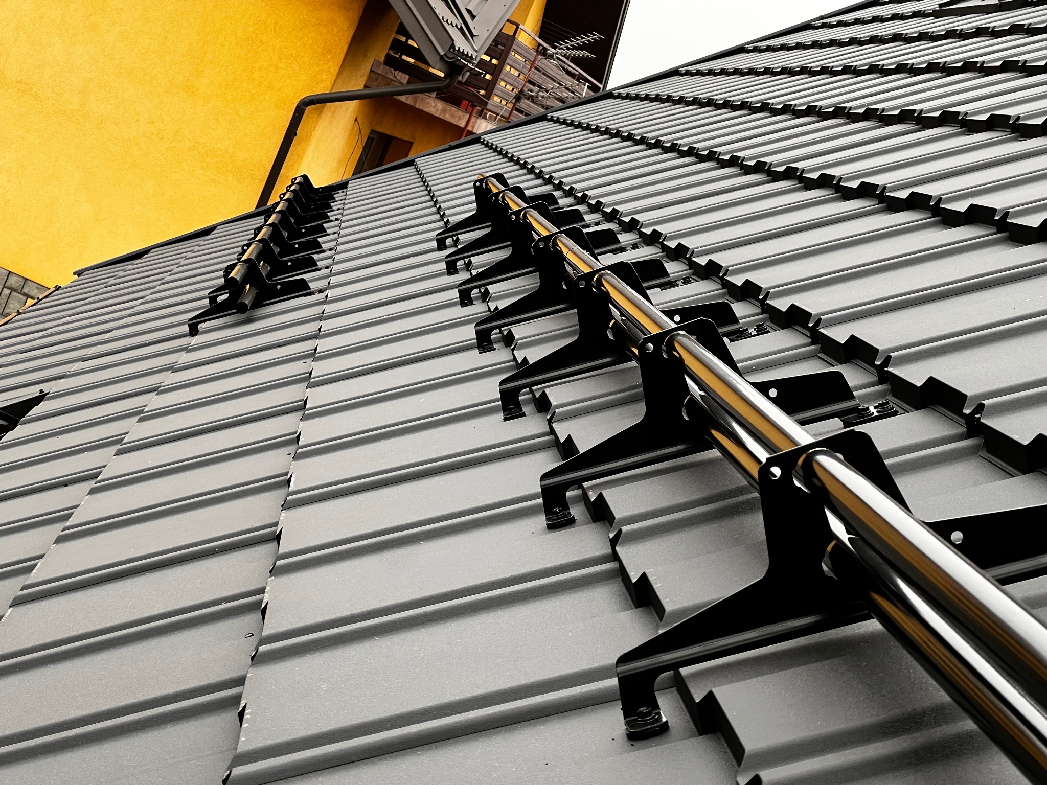 Szary dach z zamontowanymi czarnymi stopniami i rurami przeciwśniegowymi, widok z dołu, fragment żółtej ściany budynku w tle.