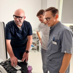 Beskid Clinic - Lekarz w okularach obsługuje aparat USG, obok dwóch obserwujących mężczyzn. Jasne wnętrze gabinetu, nowoczesny sprzęt medyczny.