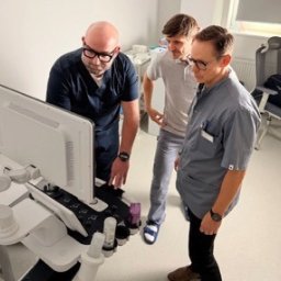Beskid Clinic - Lekarz w okularach demonstruje wyniki USG na monitorze, obok dwóch obserwujących mężczyzn w jasnym gabinecie. Sprzęt medyczny, analiza wyników badań.