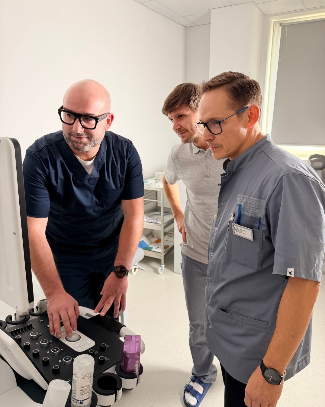 Lekarz w okularach obsługuje aparat USG, obok dwóch obserwujących mężczyzn. Jasne wnętrze gabinetu, nowoczesny sprzęt medyczny.