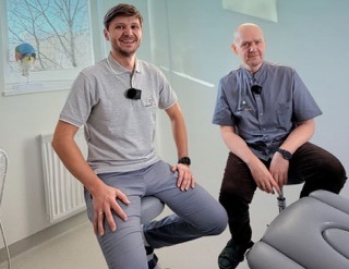 Uśmiechnięci mężczyźni w gabinecie medycznym, jeden siedzi na krześle, drugi obok. Widoczne okno i sprzęt medyczny. Obaj mają przypięte mikrofony.