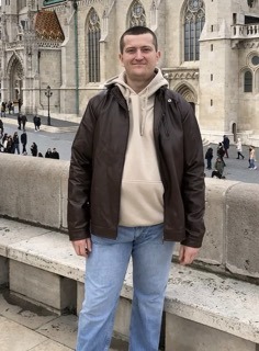 Mężczyzna w brązowej kurtce i bluzie z kapturem, na tle Bazyliki św. Macieja w Budapeszcie. Ujęcie z dołu, podkreślające architekturę i sylwetkę.