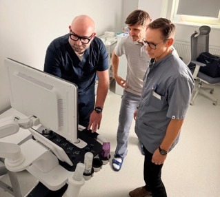 Lekarz w okularach demonstruje wyniki USG na monitorze, obok dwóch obserwujących mężczyzn w jasnym gabinecie. Sprzęt medyczny, analiza wyników badań.