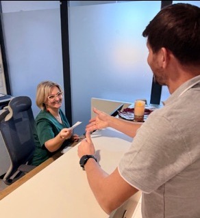 Uśmiechnięta recepcjonistka w zielonym uniformie podaje dokument pacjentowi przy jasnej ladzie recepcyjnej w nowoczesnej klinice. Pacjent wskazuje palcem na dokument.