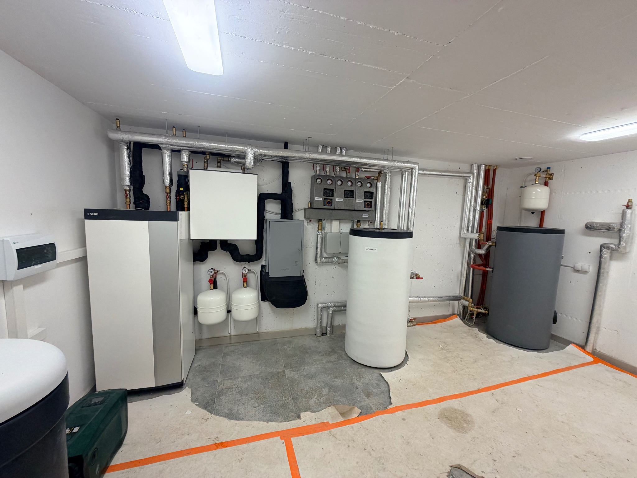 Kotłownia z zainstalowanymi urządzeniami: pompa ciepła, zbiorniki buforowe, orurowanie. Widoczne elementy instalacji grzewczej i hydraulicznej w pomieszczeniu.