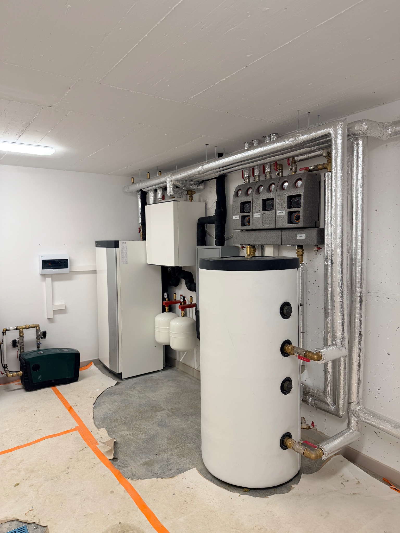 Biały zasobnik ciepłej wody z izolowanymi rurami w kotłowni. Widoczne elementy instalacji grzewczej i pompy ciepła. Podłoga z pomarańczową taśmą.