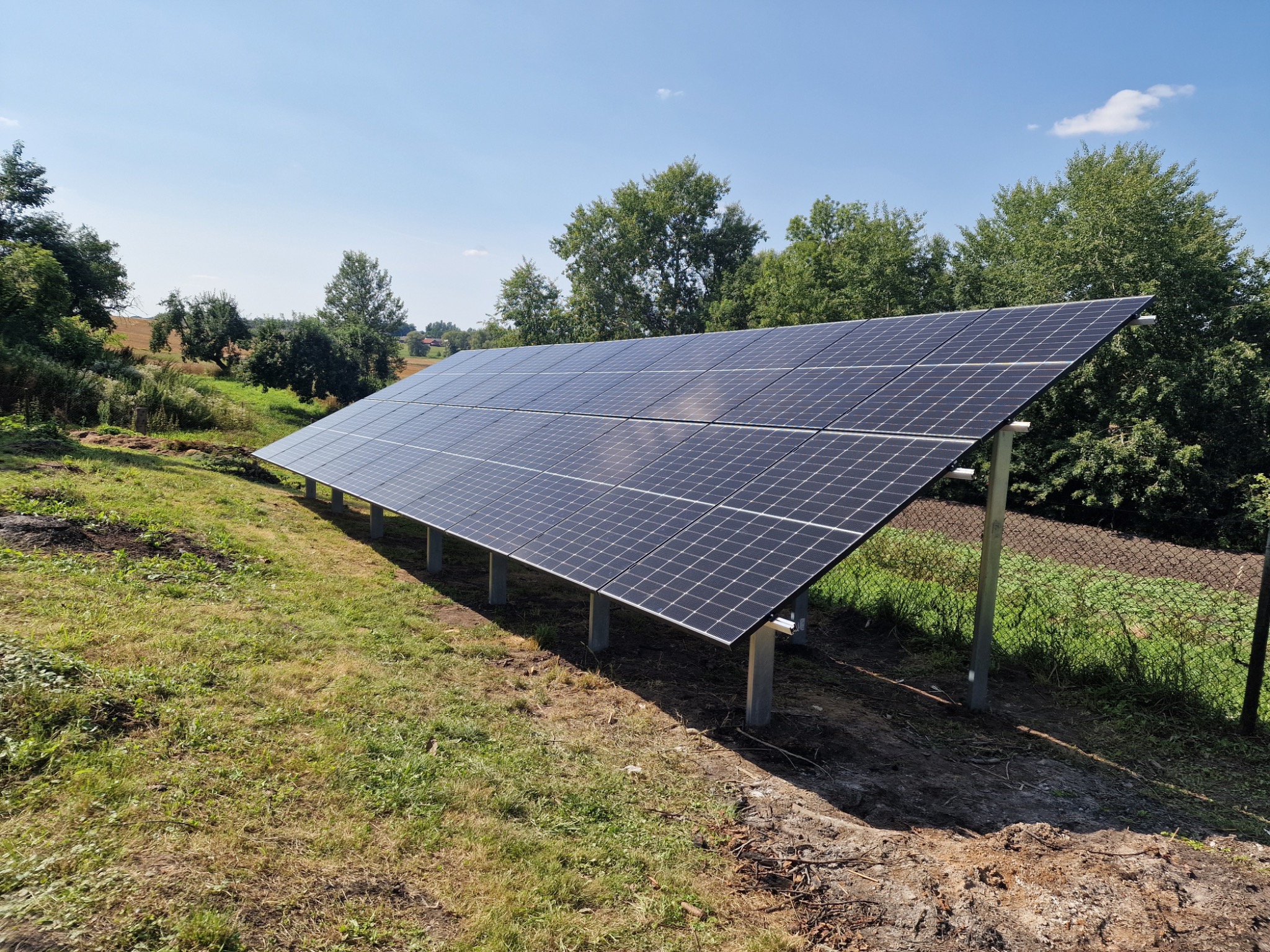 Panel słoneczny na trawiastym terenie, widok z dołu, błękitne niebo w tle. Instalacja fotowoltaiczna z widocznymi podporami i fragmentem ogrodzenia.