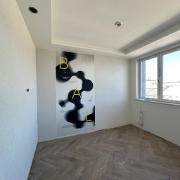 VLADYSLAV MISKIV V-LEGACY - Pokój po remoncie: parkiet w jodełkę, gładkie, białe ściany z plakatem, okno z widokiem na miasto. Widoczne ślady po gniazdkach elektrycznych i otwór na oświetlenie.