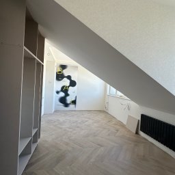 VLADYSLAV MISKIV V-LEGACY - Podłoga ułożona w jodełkę w pokoju na poddaszu, widoczny regał z półkami, ściany przygotowane do malowania, grzejnik i okno w tle.