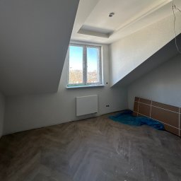 VLADYSLAV MISKIV V-LEGACY - Pokój na poddaszu w Krakowie po remoncie: świeżo pomalowane ściany, ułożona podłoga w jodełkę, okno z widokiem na miasto. Widoczne zabezpieczenia podłogi.