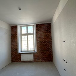 VLADYSLAV MISKIV V-LEGACY - Pokój po szpachlowaniu ścian, z fragmentem ściany z cegły. Widoczne gniazdka elektryczne i okno z grzejnikiem. Podłoga wylewka samopoziomująca.