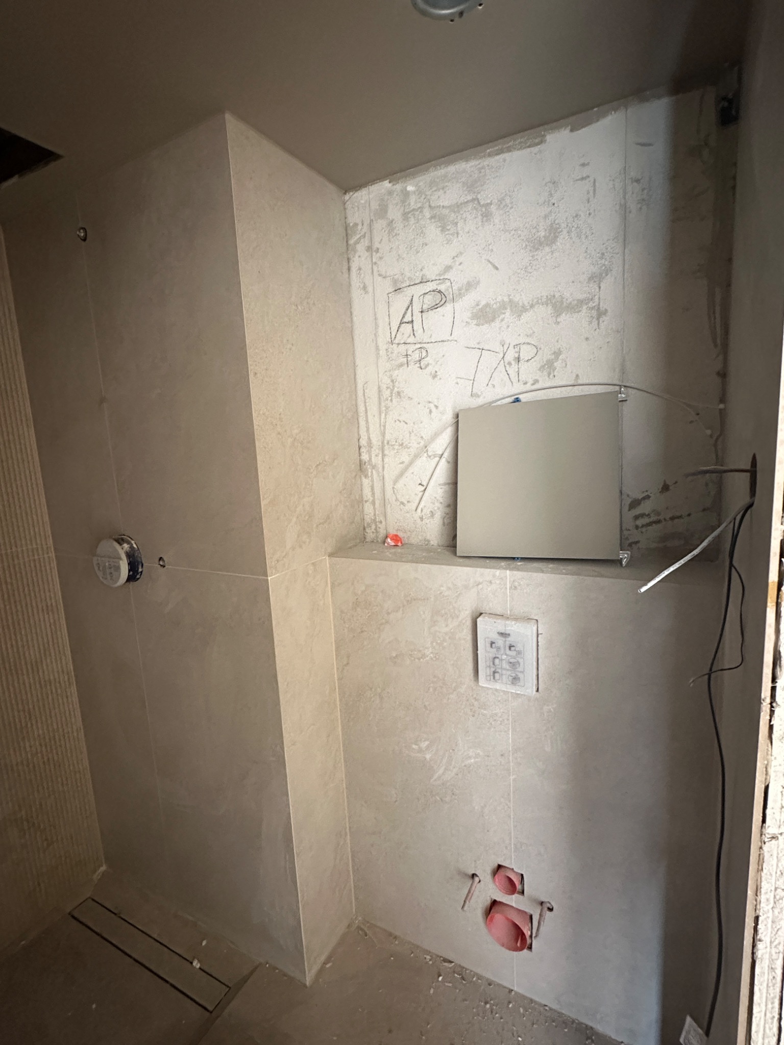 Wnętrze łazienki w trakcie wykańczania: ułożone płytki ścienne w odcieniach beżu, widoczne instalacje, fragment ściany z surowym tynkiem i odręcznymi napisami.