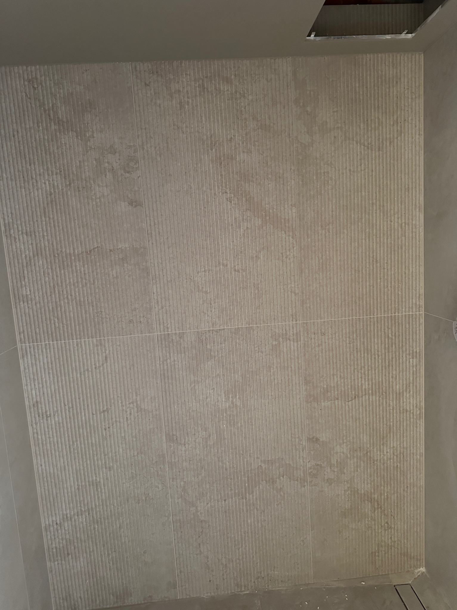 Beżowe, pionowo żłobkowane płytki ścienne w łazience, ułożone z precyzją. Widoczny fragment sufitu z otworem. Minimalistyczny design, ciepła kolorystyka.