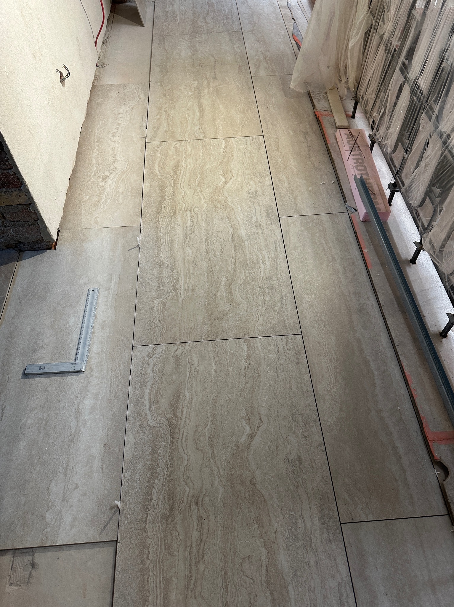 Ułożone beżowe płytki podłogowe z wzorem imitującym kamień, widoczna poziomica i krzyżyki dystansowe. Częściowo widoczne profile i folia ochronna.