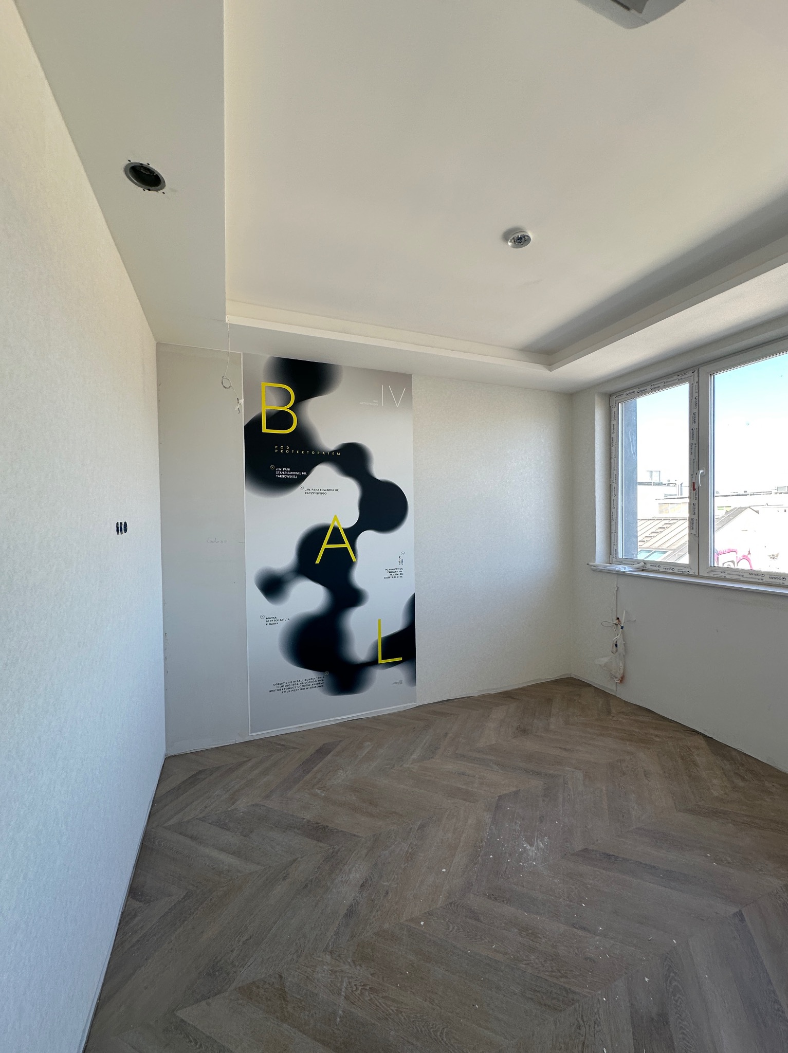 Pokój po remoncie: parkiet w jodełkę, gładkie, białe ściany z plakatem, okno z widokiem na miasto. Widoczne ślady po gniazdkach elektrycznych i otwór na oświetlenie.