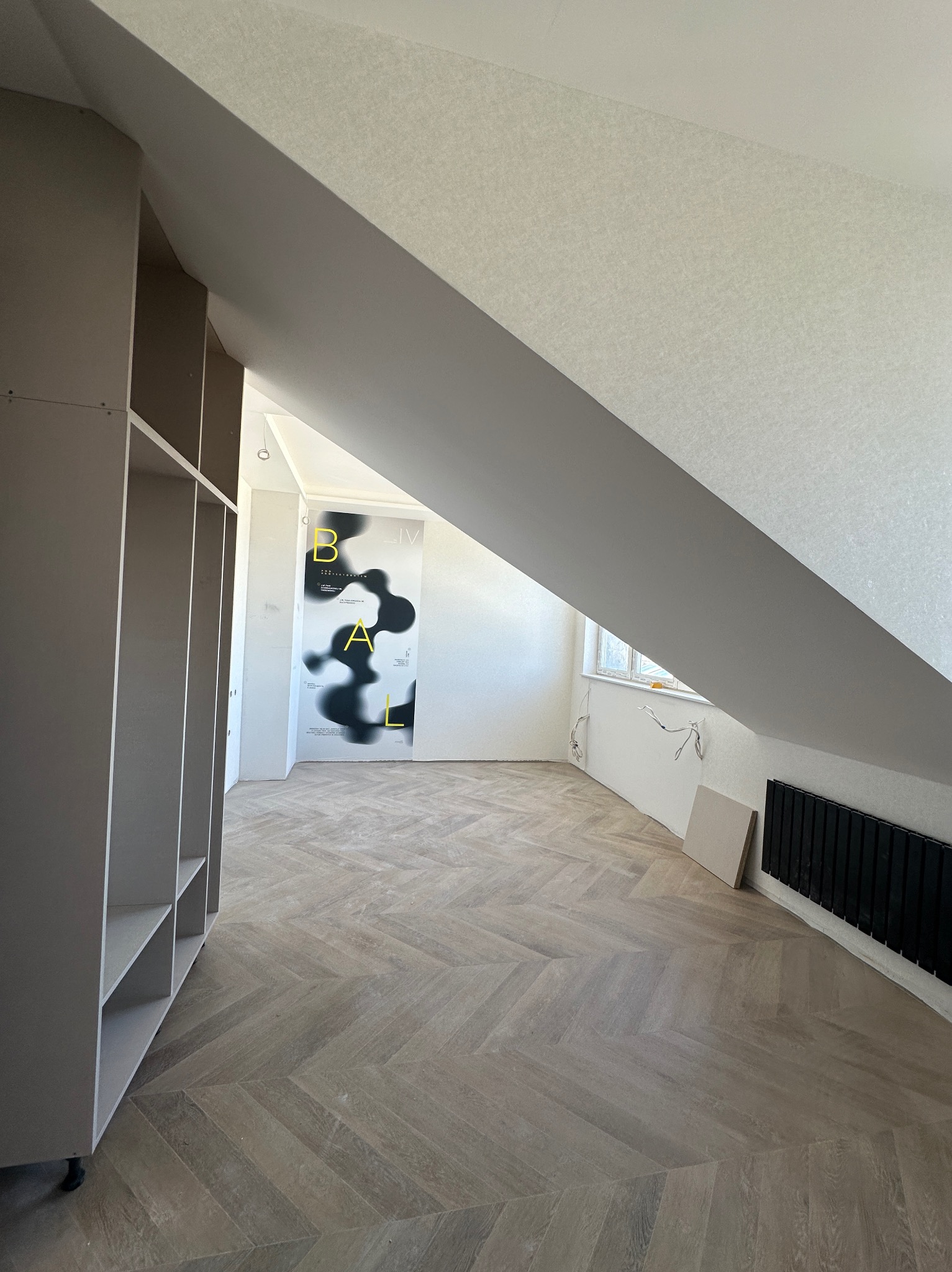 Podłoga ułożona w jodełkę w pokoju na poddaszu, widoczny regał z półkami, ściany przygotowane do malowania, grzejnik i okno w tle.