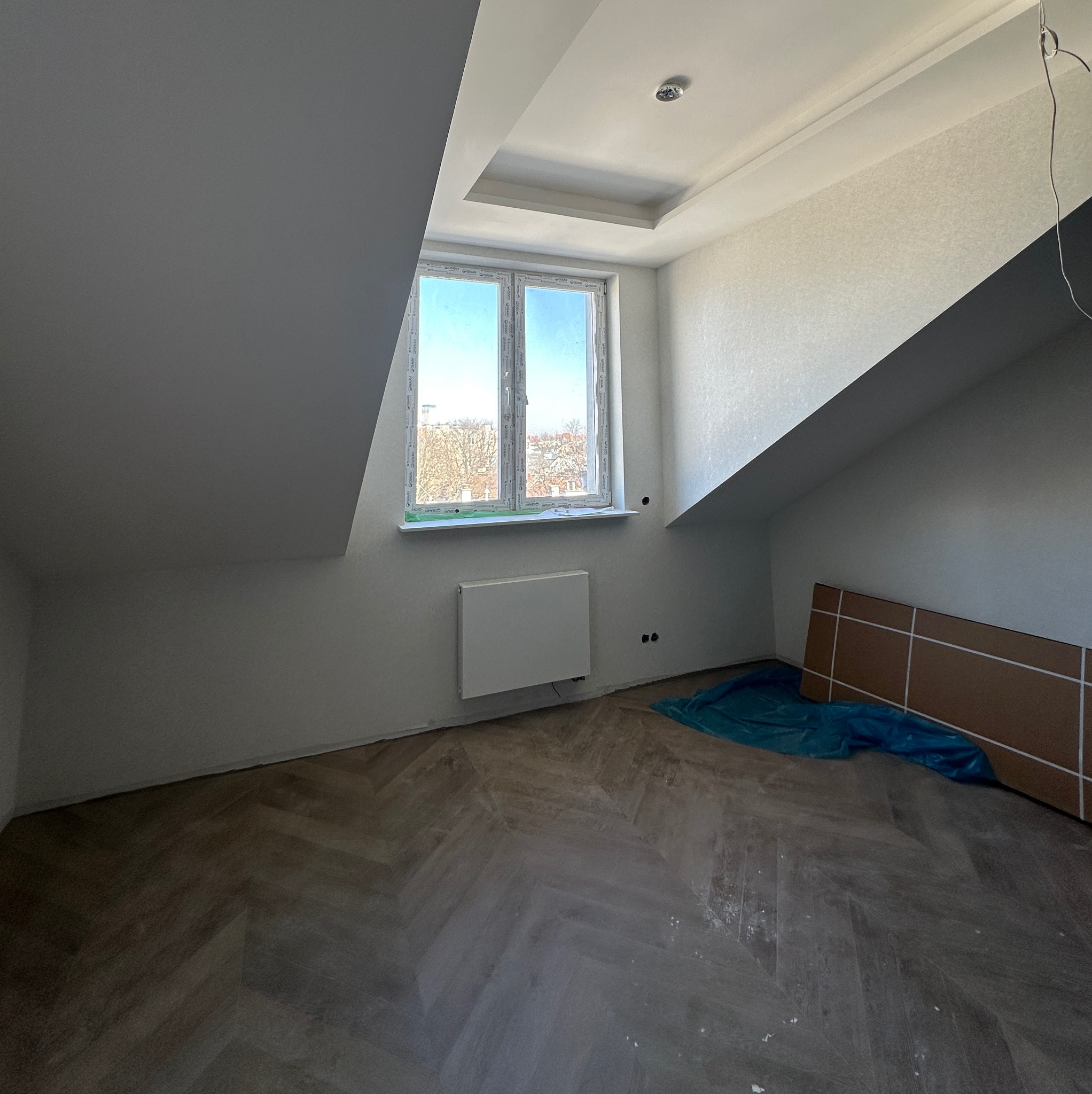 Pokój na poddaszu w Krakowie po remoncie: świeżo pomalowane ściany, ułożona podłoga w jodełkę, okno z widokiem na miasto. Widoczne zabezpieczenia podłogi.