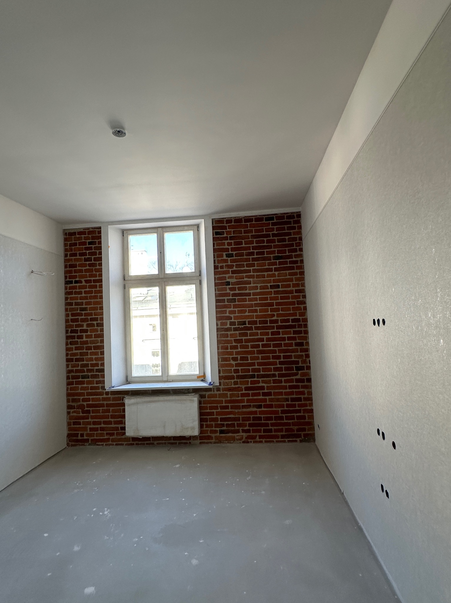 Pokój po szpachlowaniu ścian, z fragmentem ściany z cegły. Widoczne gniazdka elektryczne i okno z grzejnikiem. Podłoga wylewka samopoziomująca.