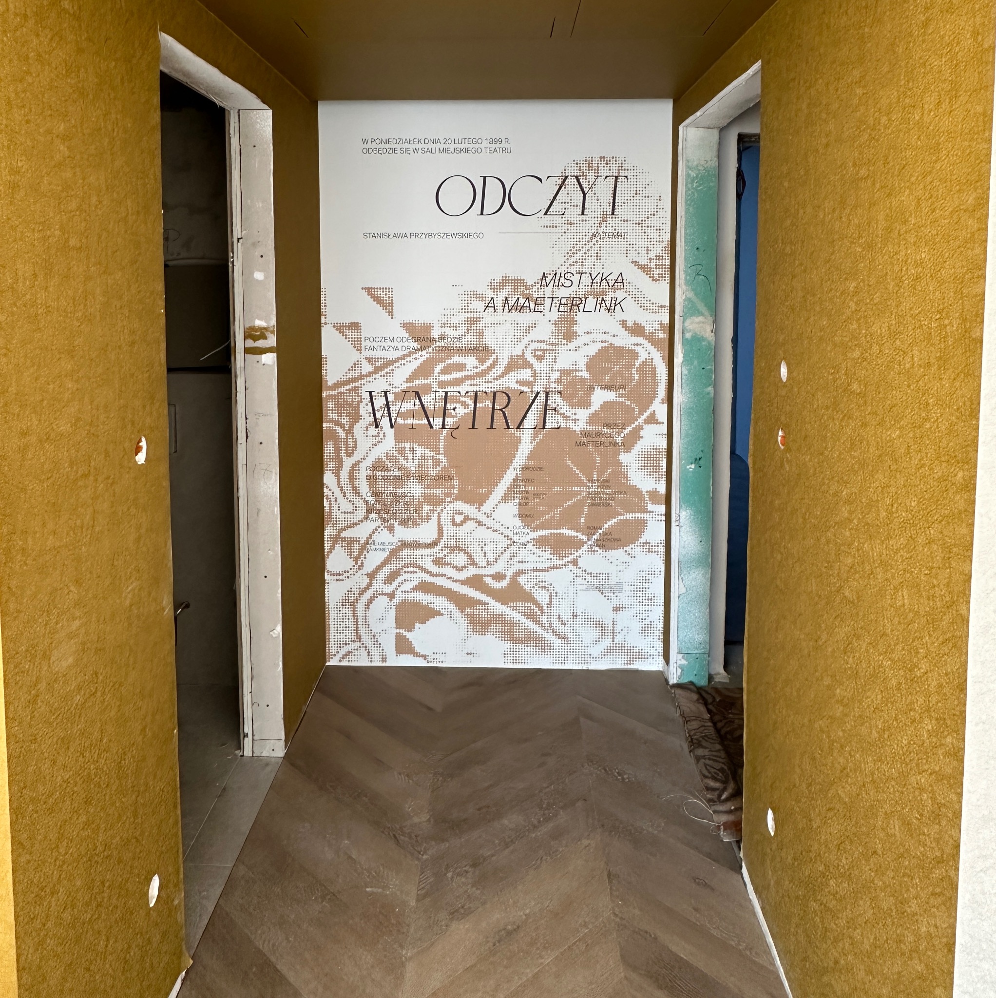 Korytarz po remoncie z nową podłogą w jodełkę i ścianami w kolorze musztardowym. Na ścianie plakat z napisem 'Wnętrze'. Widoczne niedokończone obramowania drzwi.
