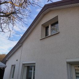 Elewacja domu jednorodzinnego z oknami i brązowym wykończeniem dachu, widziana pod kątem. Na pierwszym planie gałęzie drzewa na tle błękitnego nieba.