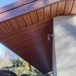 Podbitka dachowa z paneli drewnopodobnych w kolorze brązowym, zamontowana przy ścianie budynku z oświetleniem zewnętrznym. Widok z dołu na tle zieleni.